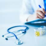 Woman's hands writing on sheet of paper on a clipboard with pen along with two vials of medication and a stethoscope illustrating opioid agonist therapy.