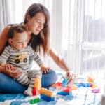 Caregiver and baby toddler playing building with learning toy stacking blocks illustrates building resilience in children.