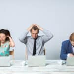 A photograph of three young professionals, each with a computer and a cup of coffee, showing signs of displacement due to stress, including grooming, lip-twisting and stretching.