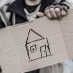 Photo of a possibly homeless person holding up a cardboard sign with a crude drawing of a house on it, symbolized the loss in household savings due to sports betting.