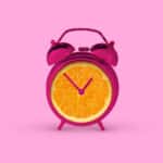 Pink alarm clock shaped like slice of orange against vibrant background symbolic of "a clockwork orange."