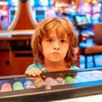 Photo of a sad child in a gambling casino standing with his hand on a box of colored tokens or chips.