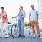 Photo of three teenagers standing in the street, two with bicycles and one with a skateboard, symbolizing exercise and community.