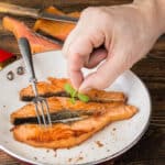 Photograph of fried salmon fillets in a pan with a hand adding garnish.