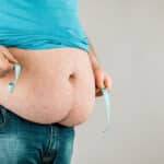 An obese man uses a measuring tape to measure his belly.