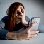 Photo of a young woman exhibiting signs of smartphone addiction holding a smartphone with her eyes closed and her hand over her face.