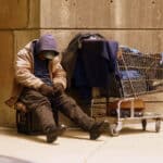 photo of a homeless person sitting on a crate next to a shopping cart full of possessions, bundled up against the freeze in funding for addiction treatment, mental health services, and services for the homeless.