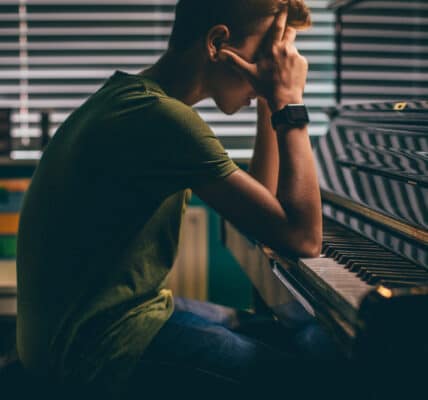 Photo of college student with his head in his hands and his elbows on an upright piano illustrating the stress cause by an addiction to playing music.