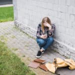 Photo of teenager sitting near wall with head in one hand and smartphone in the other, a backpack and books are strewn on the ground.