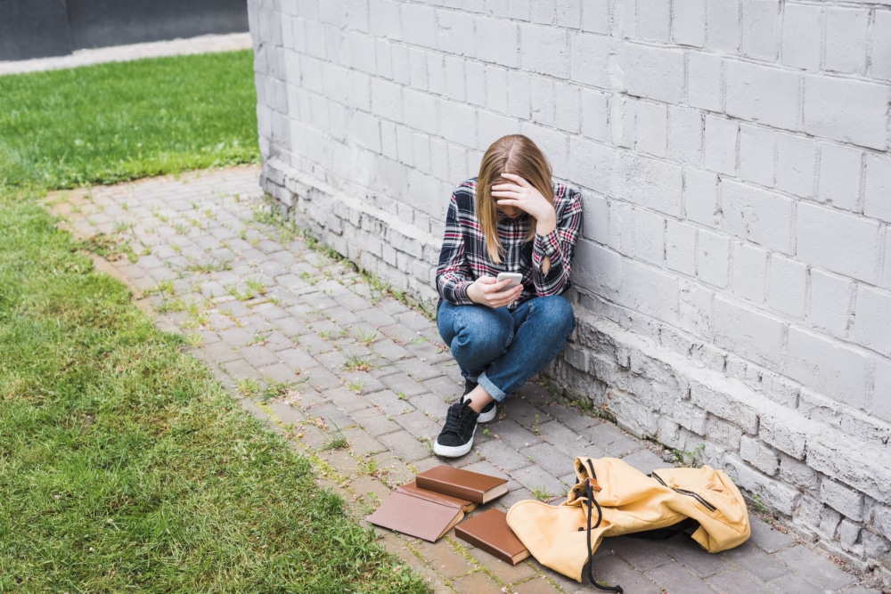 Photo of teenager sitting near wall with head in one hand and smartphone in the other, a backpack and books are strewn on the ground.