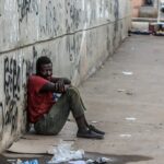 Photo of a poor man in soiled clothing sitting against a concrete wall on a filthy street illustrating some of the social components of addiction.