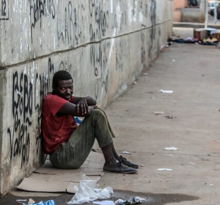 Photo of a poor man in soiled clothing sitting against a concrete wall on a filthy street illustrating some of the social components of addiction.