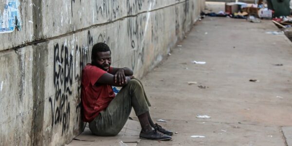 Photo of a poor man in soiled clothing sitting against a concrete wall on a filthy street illustrating some of the social components of addiction.