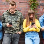 Photo of two young couples leaning against a brick wall, each of them looking at their smartphone and not interacting with each other.