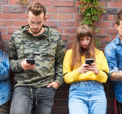 Photo of two young couples leaning against a brick wall, each of them looking at their smartphone and not interacting with each other.