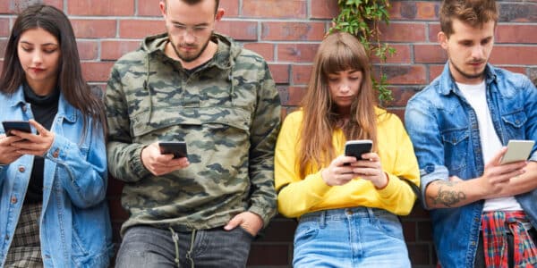 Photo of two young couples leaning against a brick wall, each of them looking at their smartphone and not interacting with each other.