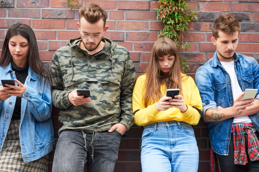 Photo of two young couples leaning against a brick wall, each of them looking at their smartphone and not interacting with each other.
