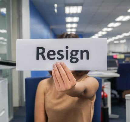 Photo of woman in an office holding up a sign with the word, "Resign."