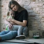 Photo of a homeless woman and her little daughter sitting near a brick wall and asking for help.