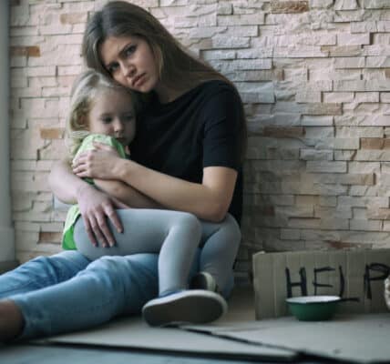 Photo of a homeless woman and her little daughter sitting near a brick wall and asking for help.