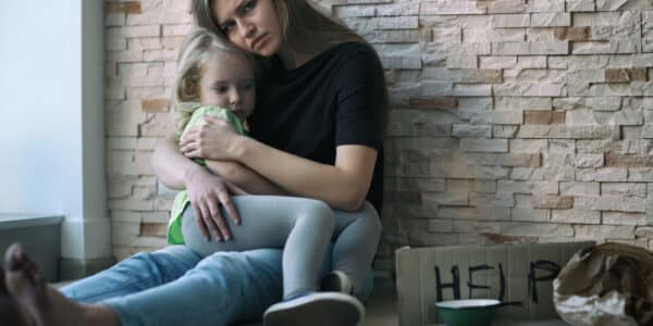 Photo of a homeless woman and her little daughter sitting near a brick wall and asking for help.