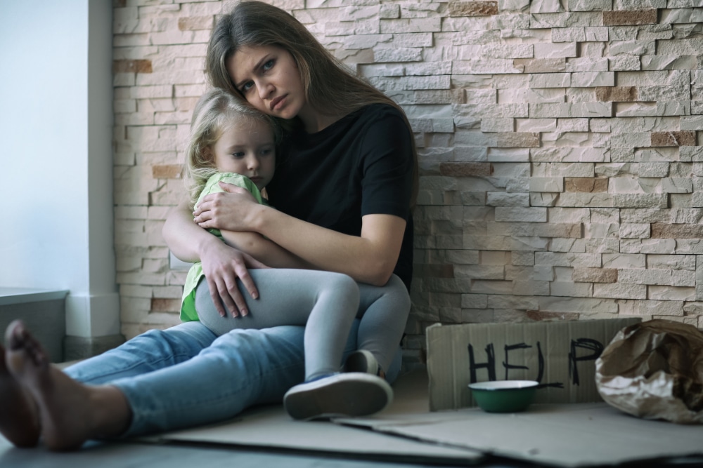 Photo of a homeless woman and her little daughter sitting near a brick wall and asking for help.