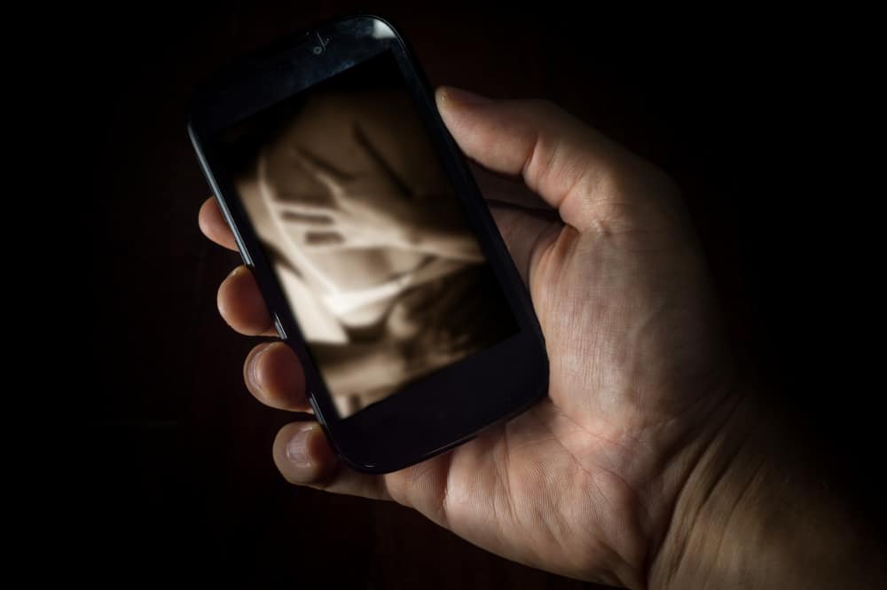 Photo of a young man's hand holding a smartphone displaying an erotic embrace.