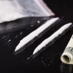 Photograph of cocaine on a table in a bag and in two lines next to a rolled-up one hundred dollar bill.