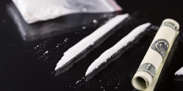 Photograph of cocaine on a table in a bag and in two lines next to a rolled-up one hundred dollar bill.
