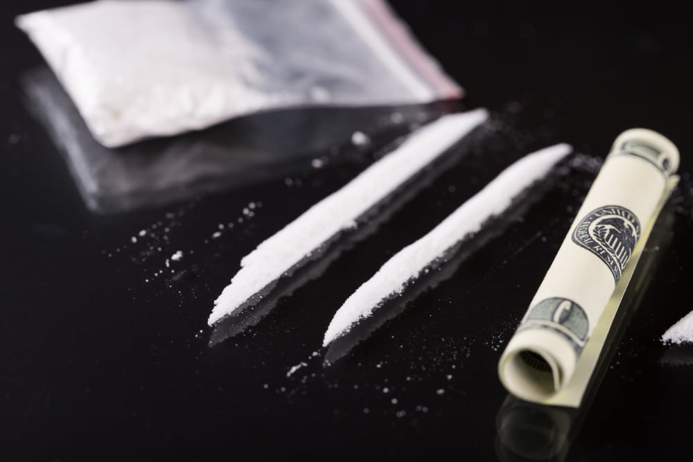 Photograph of cocaine on a table in a bag and in two lines next to a rolled-up one hundred dollar bill.
