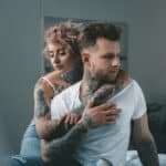 Photograph of a young man being embraced by a young woman, both heavily tattooed.