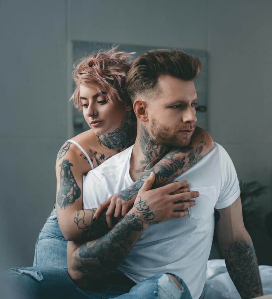 Photograph of a young man being embraced by a young woman, both heavily tattooed.