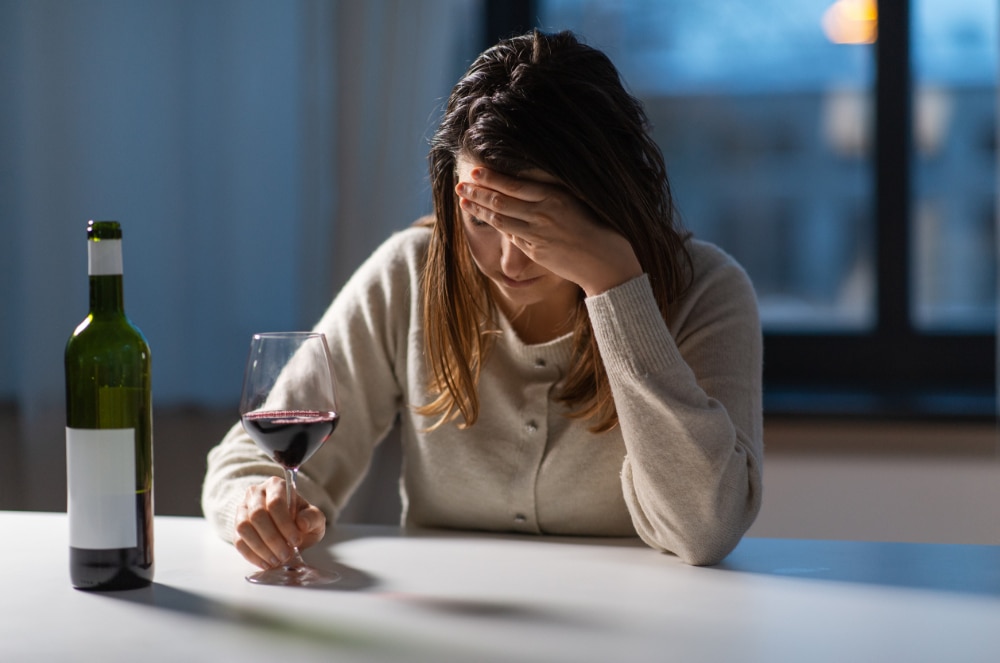 Photograph of a depressed mother with her head in her hand drinking wine.
