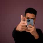 Photograph of a man interacting with an AI chatbot on a smartphone phone in his left hand while making a "finger gun" with his right hand.