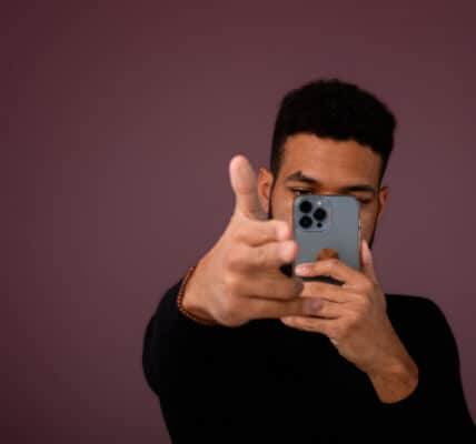 Photograph of a man interacting with an AI chatbot on a smartphone phone in his left hand while making a "finger gun" with his right hand.