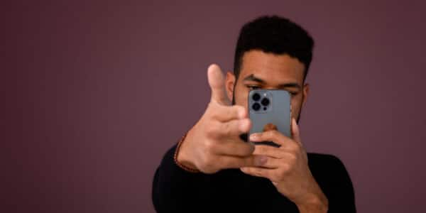 Photograph of a man interacting with an AI chatbot on a smartphone phone in his left hand while making a "finger gun" with his right hand.