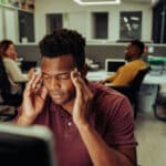Photograph of a man in an office holding his hands to his temples to indicate he is experiencing stress.