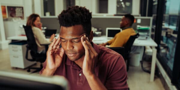 Photograph of a man in an office holding his hands to his temples to indicate he is experiencing stress.