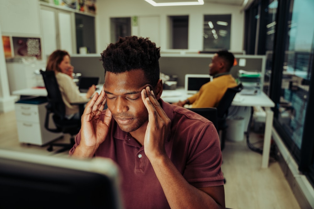 Photograph of a man in an office holding his hands to his temples to indicate he is experiencing stress.