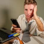 attractive sad and desperate man in lose necktie looking messy and depressed working at laptop computer desk in business office problem and stress concept holding mobile phone frustrated