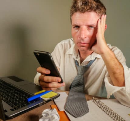 attractive sad and desperate man in lose necktie looking messy and depressed working at laptop computer desk in business office problem and stress concept holding mobile phone frustrated