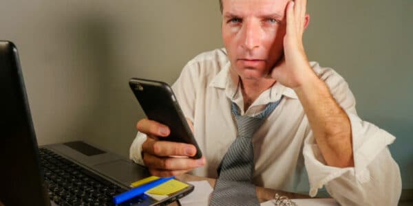 attractive sad and desperate man in lose necktie looking messy and depressed working at laptop computer desk in business office problem and stress concept holding mobile phone frustrated
