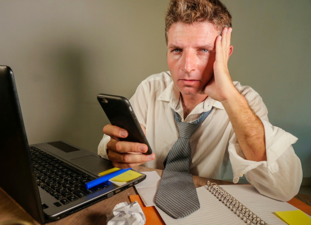 attractive sad and desperate man in lose necktie looking messy and depressed working at laptop computer desk in business office problem and stress concept holding mobile phone frustrated