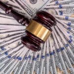 Photograph of a judge's gavel resting on top of a wheel of one hundred dollar bills.