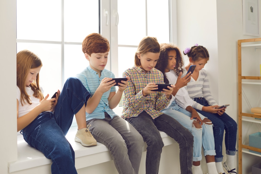 Photograph of a group of five children sitting in a window well each looking at their smartphones.