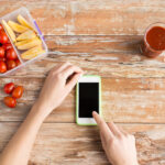 healthy eating, dieting, technology and people concept - close up of woman hands with blank smartphone and food in plastic container on table at home