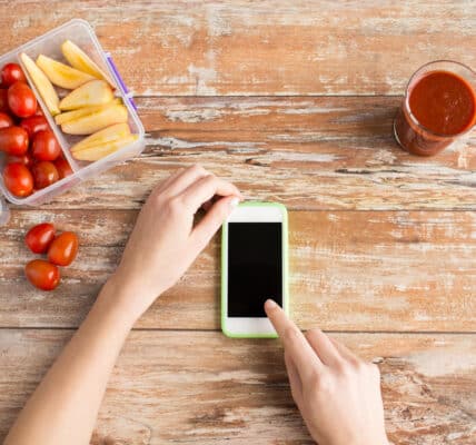 healthy eating, dieting, technology and people concept - close up of woman hands with blank smartphone and food in plastic container on table at home