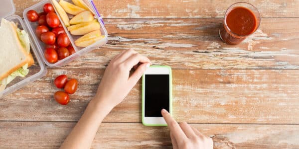 healthy eating, dieting, technology and people concept - close up of woman hands with blank smartphone and food in plastic container on table at home
