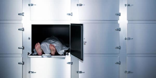 Staged photograph of a body covered with a white sheet on a morgue table in a locker.