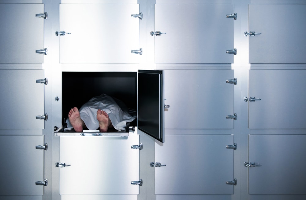 Staged photograph of a body covered with a white sheet on a morgue table in a locker.